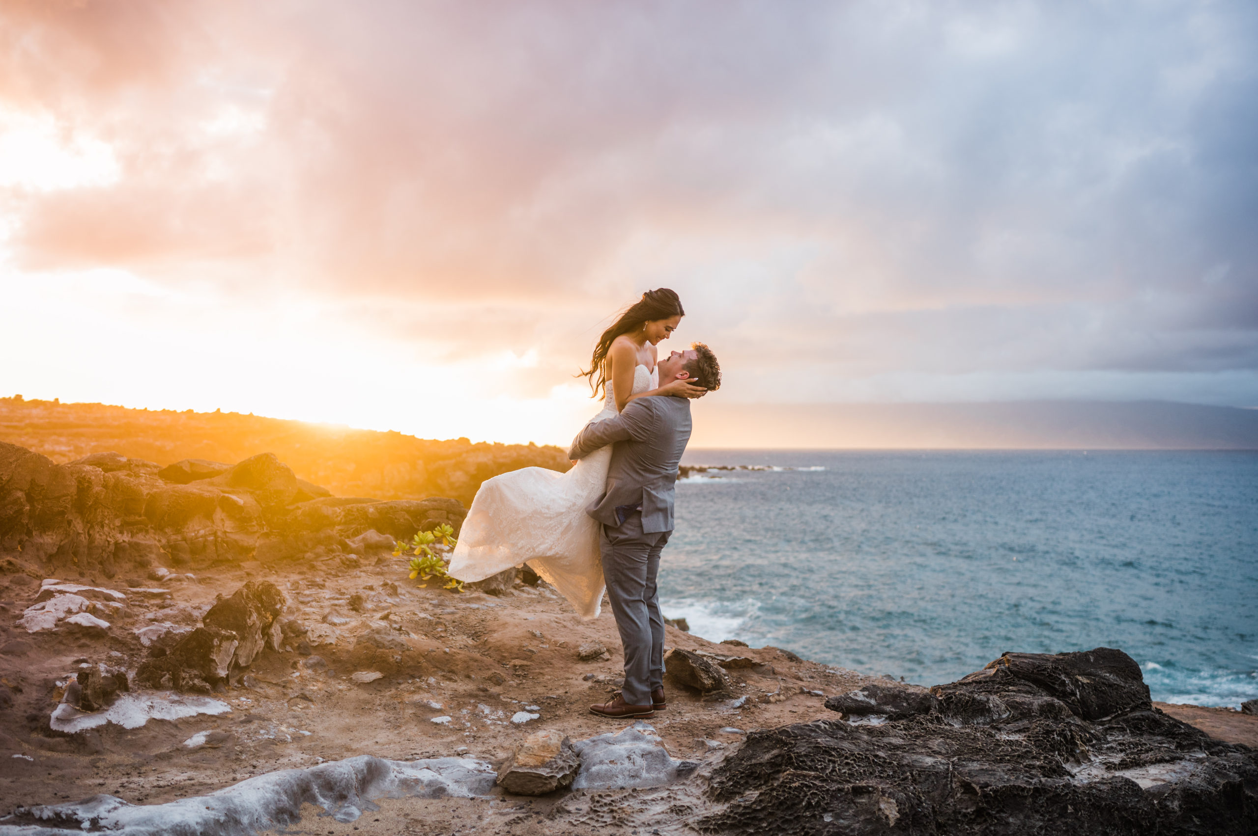 An adventurous sunset elopement at Ironwoods Cliffs in Kapalua, featuring a groom lifting his bride into the air against a backdrop of dark blue ocean waves and rugged lava rocks.