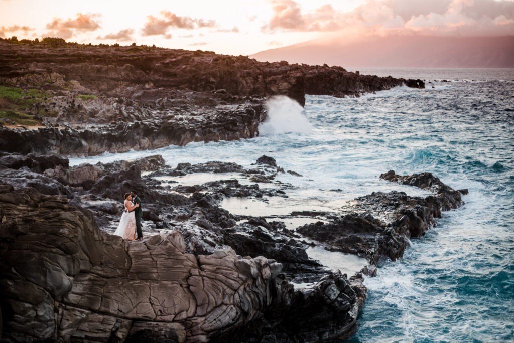 A panoramic view of an elopement at Ironwoods Cliffs in Kapalua, featuring a bride and groom embracing with rugged lava rocks, choppy waves, and the island of Molokai in the background.