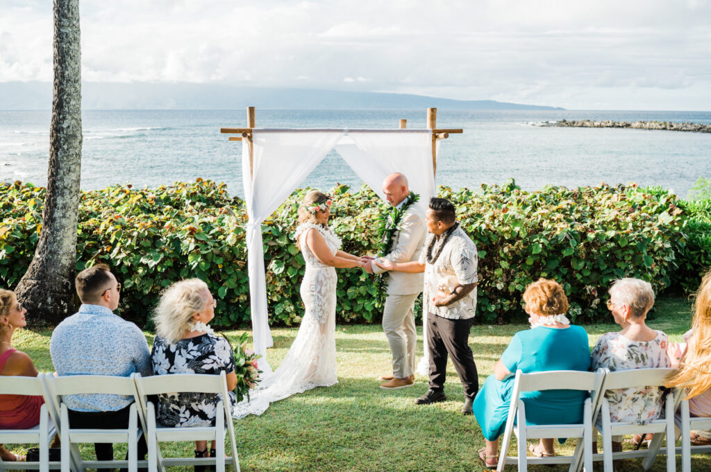 An outdoor wedding ceremony at Merriman's Kapalua, with a bride and groom holding hands in front of a calm blue ocean and wedding guests on white chairs.