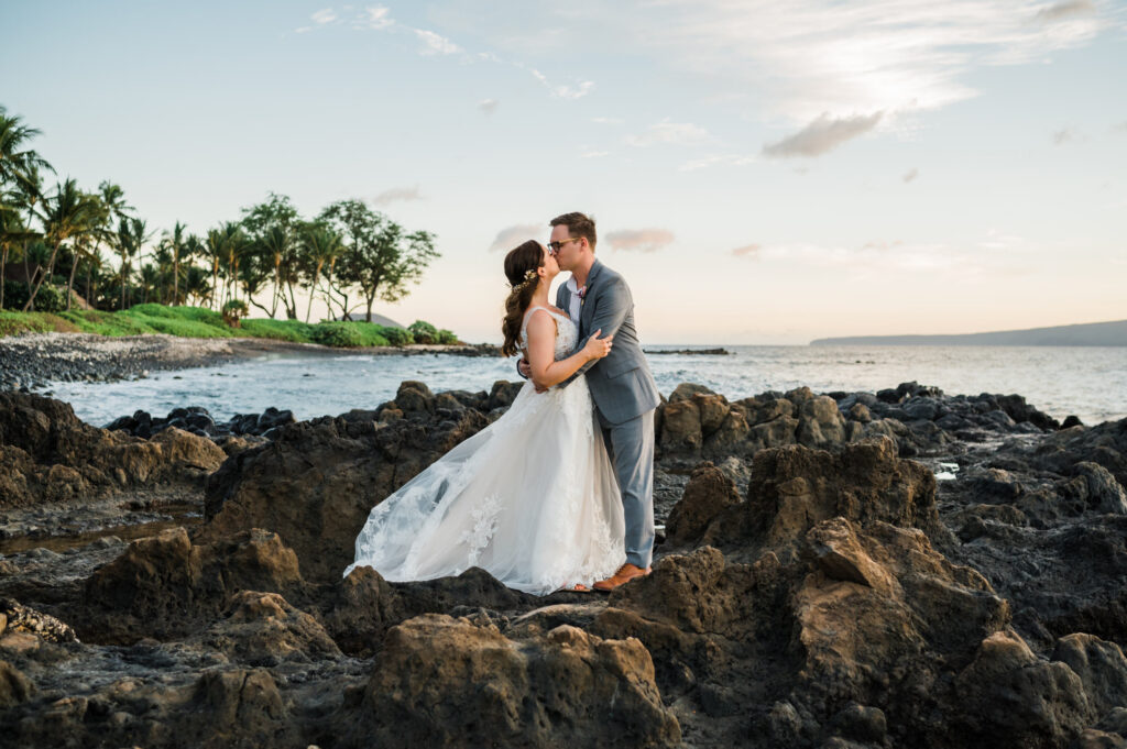 A bride and groom sharing a romantic kiss on a lava rock outcropping at Palauea Beach (White Rock Beach), with the island of Kaho‘olawe and coconut palm trees in the background.
