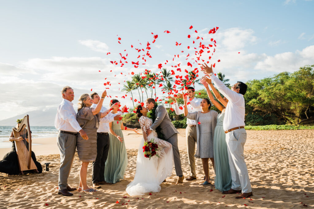 A sunny wedding celebration at Po'olenalena Beach as guests toss red flower petals around a kissing couple and a musician plays a harp in front of the ocean.