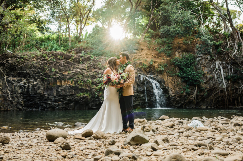 An intimate waterfall elopement at Twin Falls Maui, featuring a bride and groom gazing into each other's eyes in a lush tropical rainforest setting at sunset.