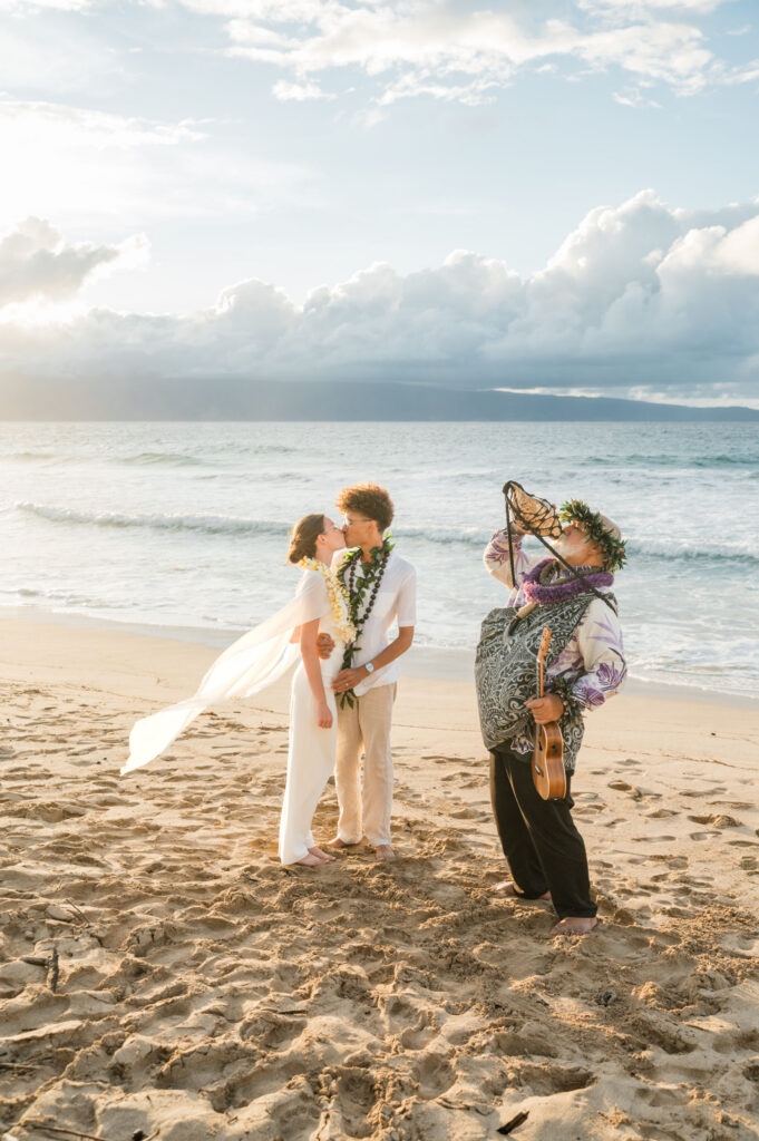 A joyful couple embraces, sharing a kiss on the sandy beach at sunset, with a musician in traditional attire playing nearby at Ironwoods Maui.