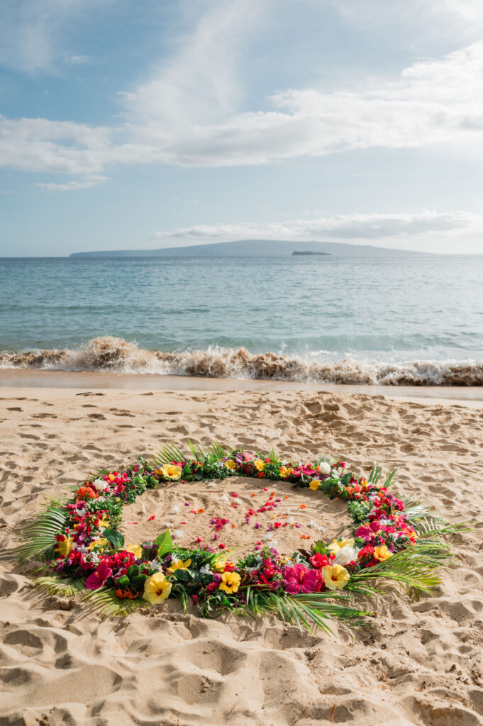 A vibrant floral circle sits in the sand, near gentle waves at Palauea beach, Maui, illuminated by soft morning light beneath a blue sky.