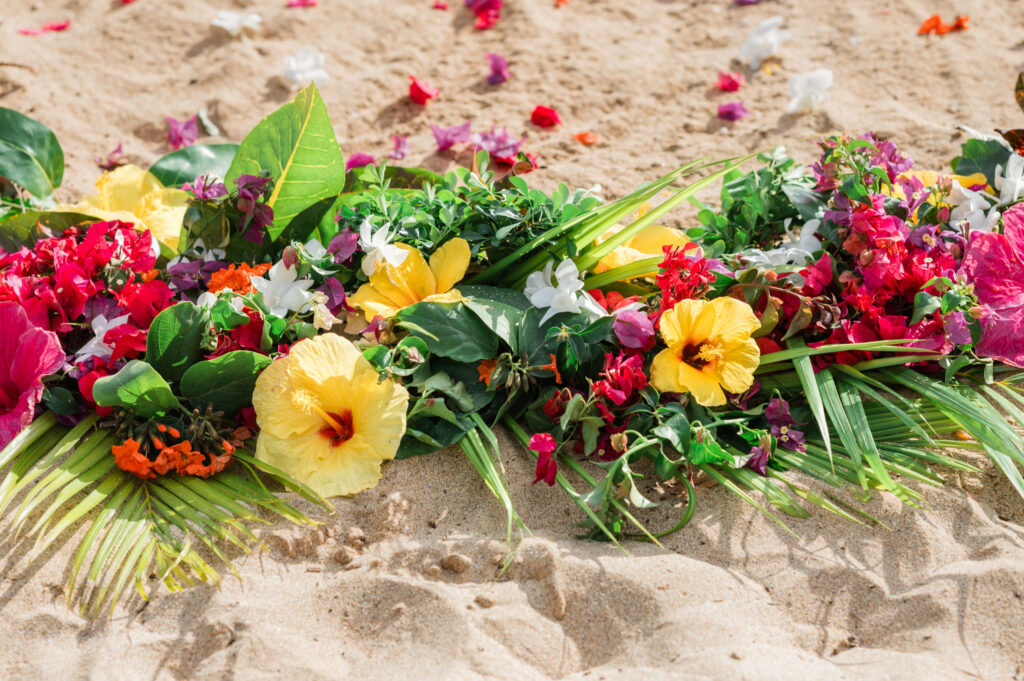 A vibrant array of tropical flowers, including hibiscus and bougainvillea, artfully arranged in the soft sand at Palauea Beach, Maui, under bright sunlight.