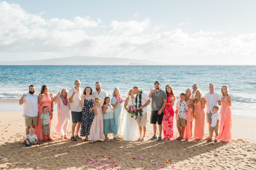 In the warm sunlight, a couple kisses at Poolenalena beach, Maui, surrounded by joyful family members in vibrant attire on the sandy shore.